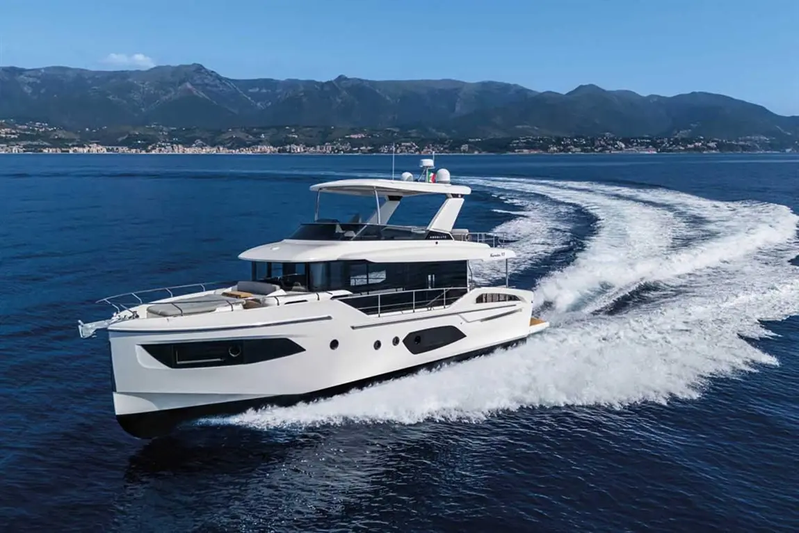 A sleek white yacht navigates smooth blue waters, leaving a wake, with mountains and a coastal town visible in the background.